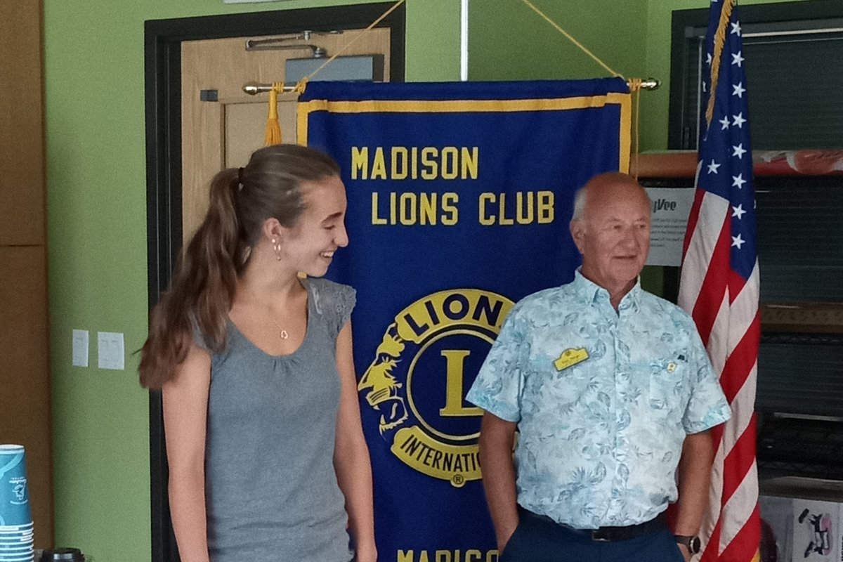 Standing in front of a Madison Lions Club banner, a man wearing a floral shirt presents a check to a young woman wearing a grey top and jeans