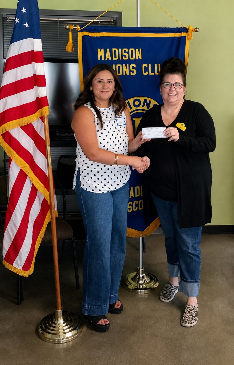 A young woman with shoulder-length dark brown hair wearing a sleeveless white blouse with black polka dots and blue jeans, shakes hands with a woman  holding a scholarship check