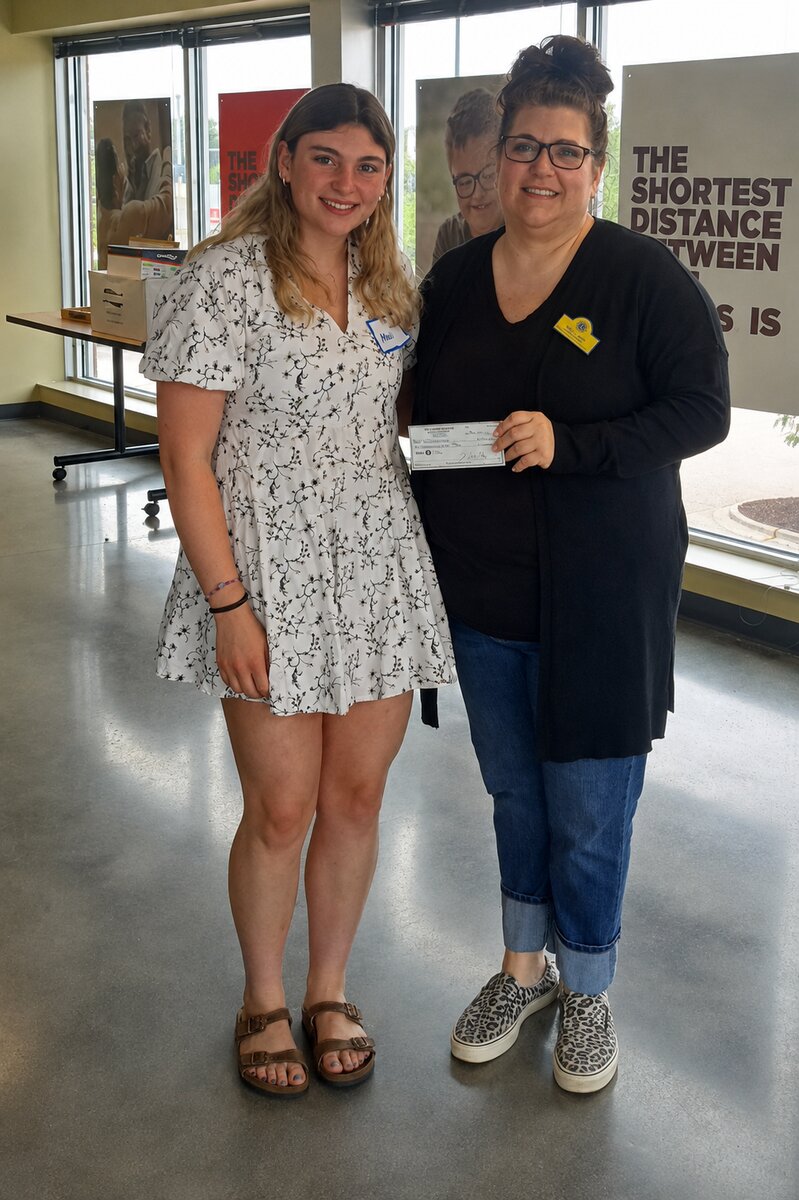A young woman with long dark-blonde hair wearing a short white dress patterned with black flowers stands beside a woman holding a scholarship check