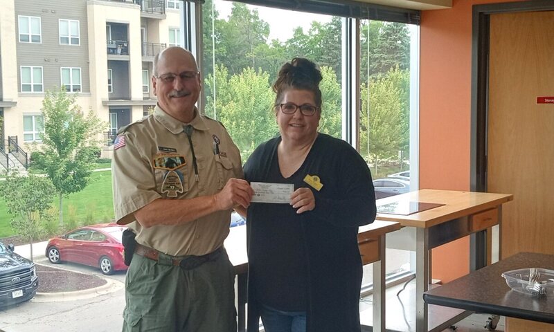 A smiling woman in glasses and a black cardigan hands a check to a mustached man in a tan scout uniform with a neckerchief.