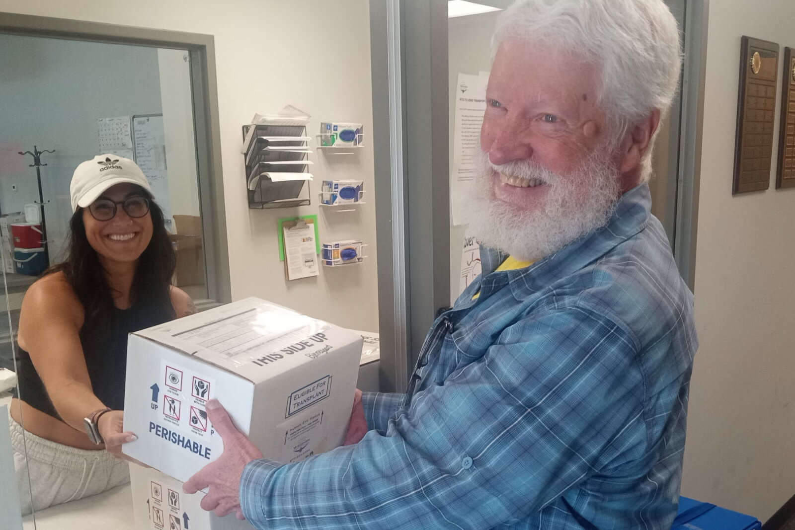 A man exchanges a box labeled “Perishable” and “Eligible for transplant” with a smiling woman behind a window.