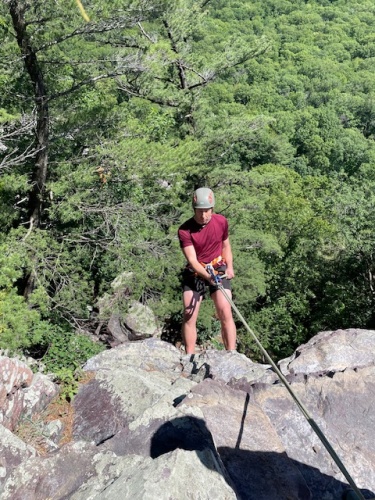 A young man wearing a climbing helmet and harness, suspended from ropes in front of a purple quartzite cliff.