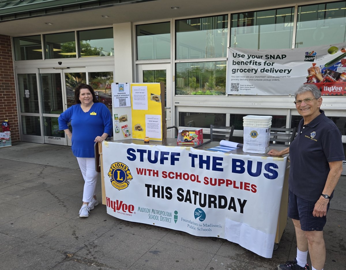 A woman in a blue shirt and a man in a navy Lions polo shirt stand behind a table with a 'Stuff the Bus' banner